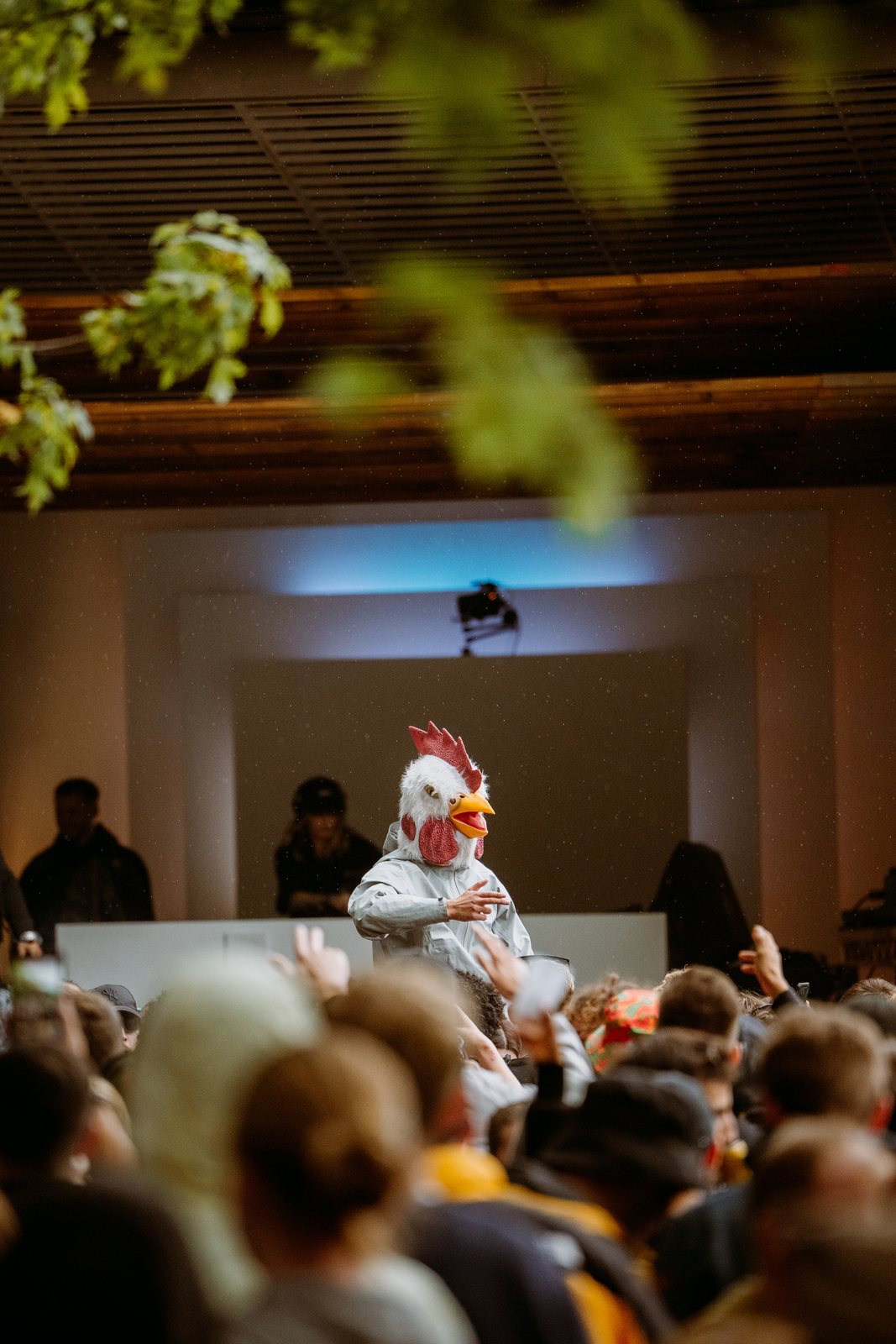 Performer in a chicken mask on stage above a packed crowd in a hall