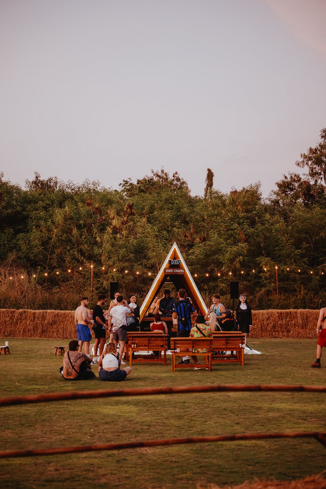 Small A-frame stage on a lawn at dusk with festoon lights and hay bales